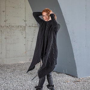 May include: A woman wearing a black knit cardigan with a long, flowing design. She is standing in front of a concrete wall and is looking to the side.