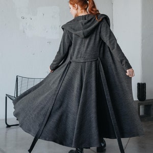 Può includere: Un lungo cappotto grigio con cappuccio e cintura da annodare. Il cappotto ha una silhouette fluida a forma di A ed è realizzato in un tessuto morbido e testurizzato.