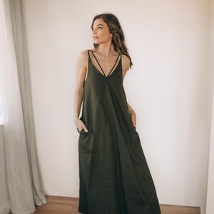 May include: Olive green maxi dress with a V-neckline and spaghetti straps. The dress features a relaxed fit with pockets and is paired with red shoes. The model is standing in front of a white wall and a window with a sheer curtain.