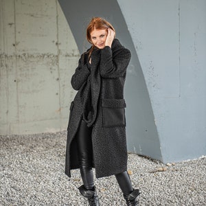 May include: A woman wearing a long, black, fuzzy coat with a large pocket on the left side. She is wearing black pants and black boots. The woman is standing in front of a concrete wall.