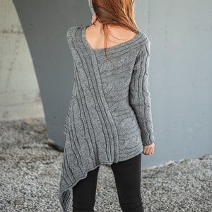 May include: A gray cable knit sweater with a draped asymmetrical hem. The sweater is made of a soft, warm material and has a relaxed fit.
