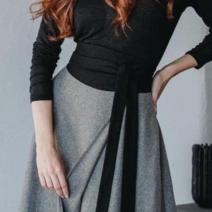 May include: A woman wearing a black long-sleeved top with a black sash tied at the waist and a gray wrap skirt. She is wearing black ankle boots.
