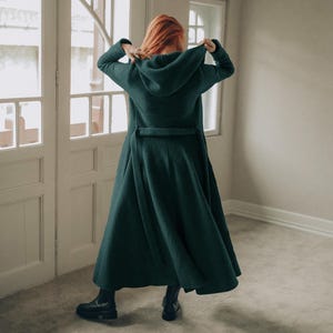 Woman wearing an elegant wool cardigan coat, standing gracefully next to a window. Handmade from a soft and cozy wool blend, this long winter coat features functional pockets, a generously sized hood, and a full-length drape for maximum comfort and style.