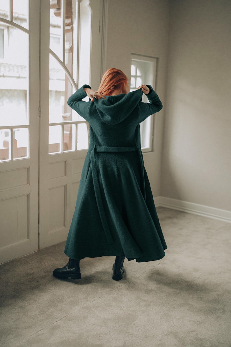 Woman wearing an elegant wool cardigan coat, standing gracefully next to a window. Handmade from a soft and cozy wool blend, this long winter coat features functional pockets, a generously sized hood, and a full-length drape for maximum comfort and style.