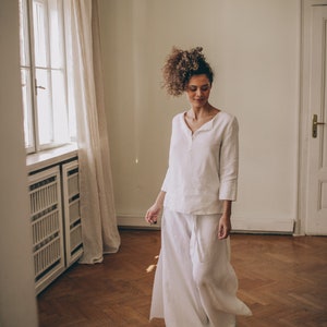 May include: A woman wearing a white linen outfit, including a long-sleeved top and wide-leg pants. The outfit has a relaxed fit and a minimalist design. The setting is a room with a wooden floor and a window with a curtain.