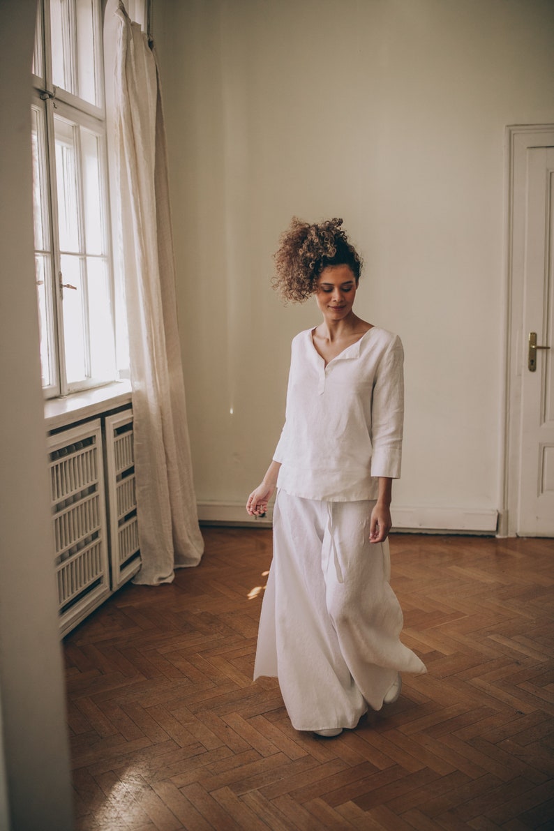 May include: A woman wearing a white linen outfit, including a long-sleeved top and wide-leg pants. The outfit has a relaxed fit and a minimalist design. The setting is a room with a wooden floor and a window with a curtain.