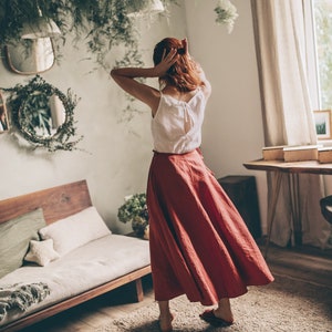 May include: A woman wearing a white top and a long, flowing, rust-colored skirt. The top has a bow detail on the back. The skirt is made of a lightweight fabric. The room has a natural, bohemian aesthetic with plants and wooden furniture.