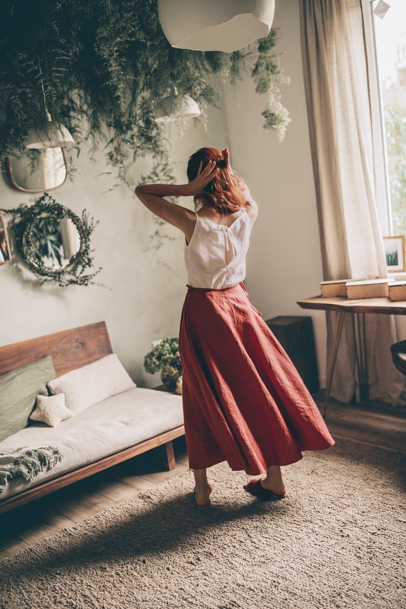 May include: A woman wearing a white top and a long, flowing, rust-colored skirt. The top has a bow detail on the back. The skirt is made of a lightweight fabric. The room has a natural, bohemian aesthetic with plants and wooden furniture.