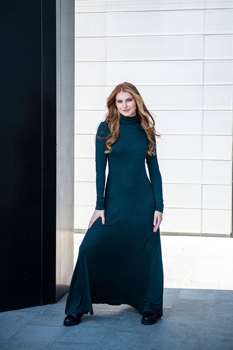 May include: A woman wearing a long, dark green, turtleneck dress with long sleeves. She is standing in front of a white wall with black trim.