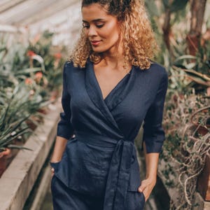 Könnte beinhalten: Ein marineblauer Wickel-Jumpsuit mit passendem Gürtel. Das Outfit hat einen V-Ausschnitt und Dreiviertelärmel. Das Model steht in einem Gewächshaus mit üppigem Grün im Hintergrund.