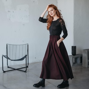 May include: A woman wearing a black long-sleeved shirt and a burgundy maxi skirt. She is standing in a room with a white wall and a black metal chair.