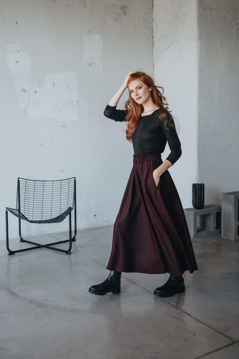 May include: A woman wearing a black long-sleeved shirt and a burgundy maxi skirt. She is standing in a room with a white wall and a black metal chair.