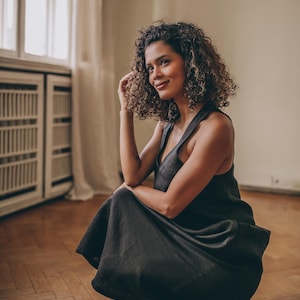 May include: A dark gray, sleeveless dress with a V-neck. The dress is worn by a person with curly hair, who is squatting on a wooden floor. The dress has a flowing skirt and a simple, elegant design. The background includes a window and a cabinet.