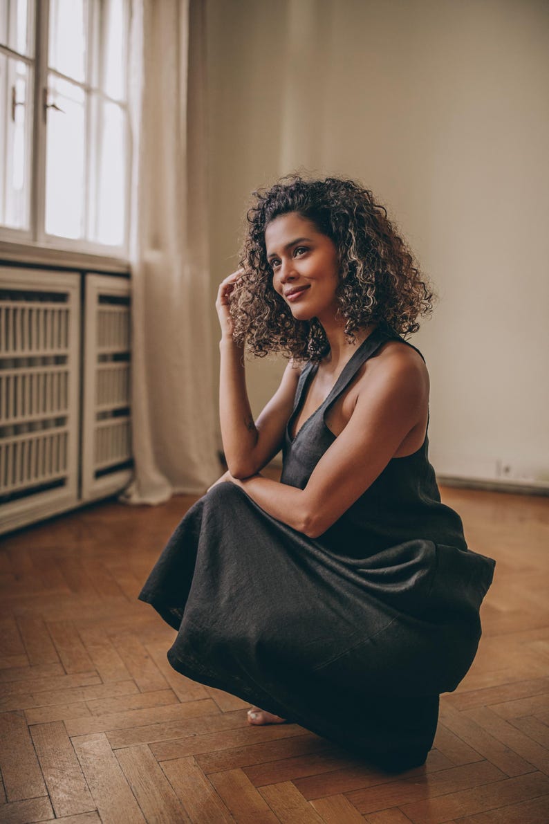 May include: A dark gray, sleeveless dress with a V-neck. The dress is worn by a person with curly hair, who is squatting on a wooden floor. The dress has a flowing skirt and a simple, elegant design. The background includes a window and a cabinet.