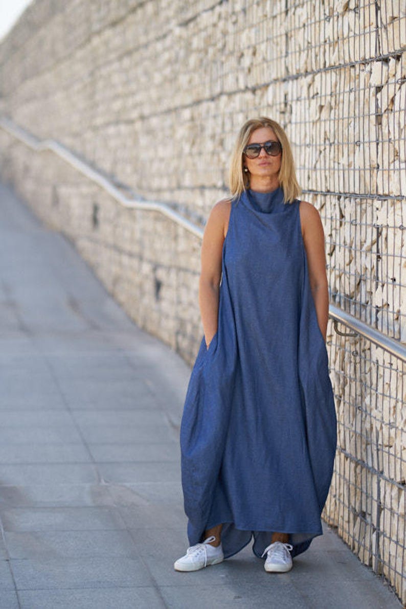 Puede incluir: Una mujer con un vestido de mezclilla azul con cuello alto y bolsillos. Lleva zapatillas blancas y gafas de sol.