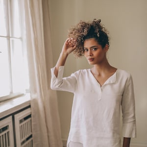 May include: White linen top and matching pants. The top has a V-neck and three-quarter sleeves with a split cuff. The pants are wide-legged. The outfit is made of a lightweight, breathable fabric, perfect for warm weather.
