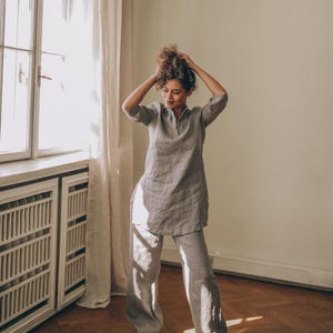 May include: A light gray linen tunic and matching wide-leg pants set. The tunic has a mandarin collar and three-quarter sleeves. The pants are loose-fitting. The outfit is worn with white sneakers. The model is standing in a room with a window and curtains.