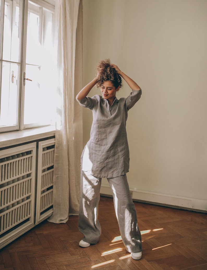 May include: A light gray linen tunic and matching wide-leg pants set. The tunic has a mandarin collar and three-quarter sleeves. The pants are loose-fitting. The outfit is worn with white sneakers. The model is standing in a room with a window and curtains.