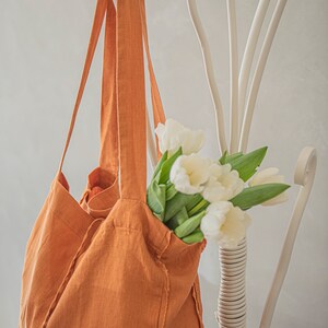 May include: A large, orange linen tote bag with frayed edges hangs on a white metal rack. A bouquet of white tulips with green stems is inside the bag.