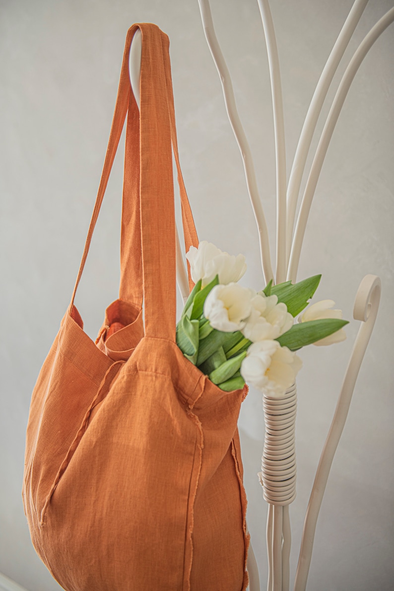May include: A large, orange linen tote bag with frayed edges hangs on a white metal rack. A bouquet of white tulips with green stems is inside the bag.