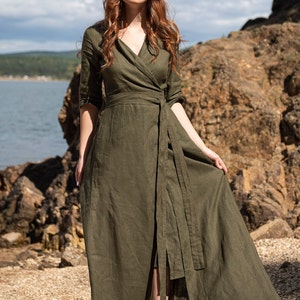 Puede incluir: Un vestido cruzado verde oliva con cuello en V y cintur&oacute;n en la cintura. El vestido tiene mangas tres cuartos y una falda larga y fluida. La modelo est&aacute; de pie al aire libre cerca de un cuerpo de agua.