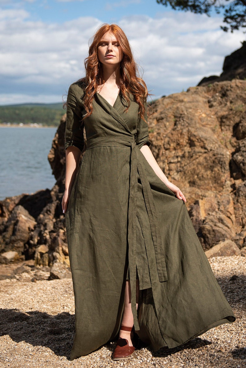 Puede incluir: Un vestido cruzado verde oliva con cuello en V y cintur&oacute;n en la cintura. El vestido tiene mangas tres cuartos y una falda larga y fluida. La modelo est&aacute; de pie al aire libre cerca de un cuerpo de agua.