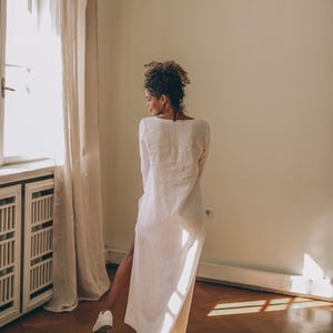 May include: A long, white linen dress with a high side slit. The dress has long sleeves and a scoop neckline. The person is wearing white sneakers. The room has a window with a beige curtain and a wooden floor.
