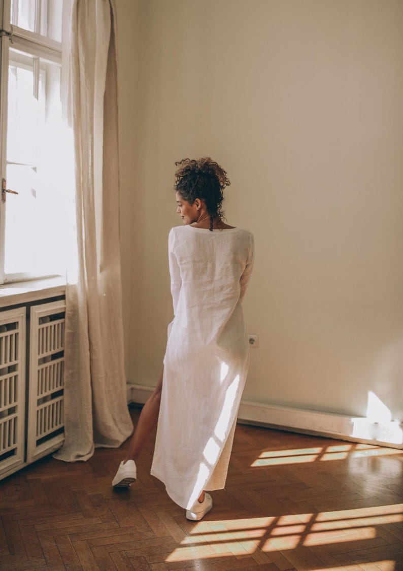May include: A long, white linen dress with a high side slit. The dress has long sleeves and a scoop neckline. The person is wearing white sneakers. The room has a window with a beige curtain and a wooden floor.