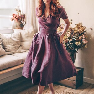 May include: A plum-colored wrap dress with short sleeves and a flowing skirt. The dress is cinched at the waist with a matching belt. The model is wearing burgundy sandals. The setting is a well-lit room with a neutral color palette.