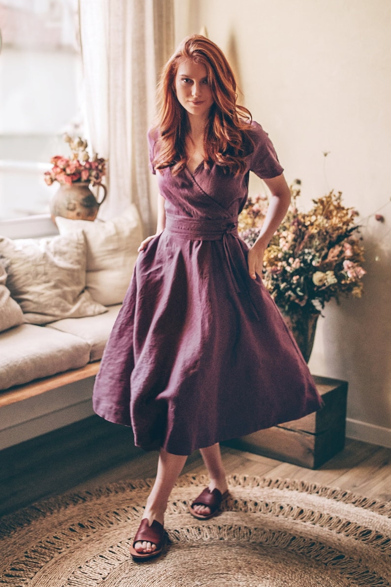 May include: A plum-colored wrap dress with short sleeves and a flowing skirt. The dress is cinched at the waist with a matching belt. The model is wearing burgundy sandals. The setting is a well-lit room with a neutral color palette.