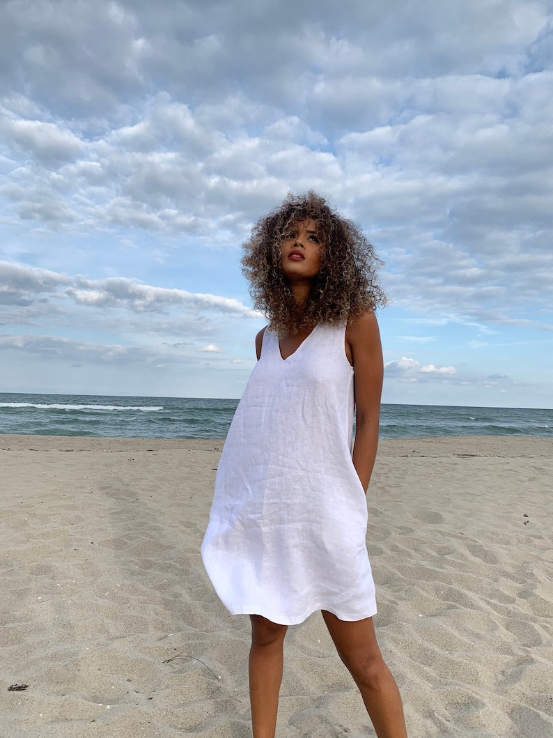May include: A woman wearing a white linen dress stands on a sandy beach with the sea in the background. The sky is cloudy and blue.