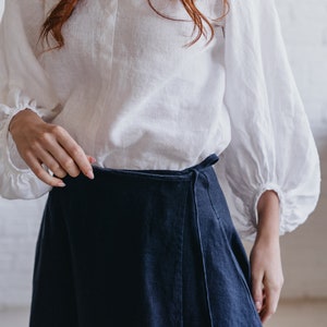 May include: A white linen blouse with long, gathered sleeves and a dark blue wrap skirt. The blouse has a simple neckline and the skirt ties at the waist. The outfit is made of natural fabrics.