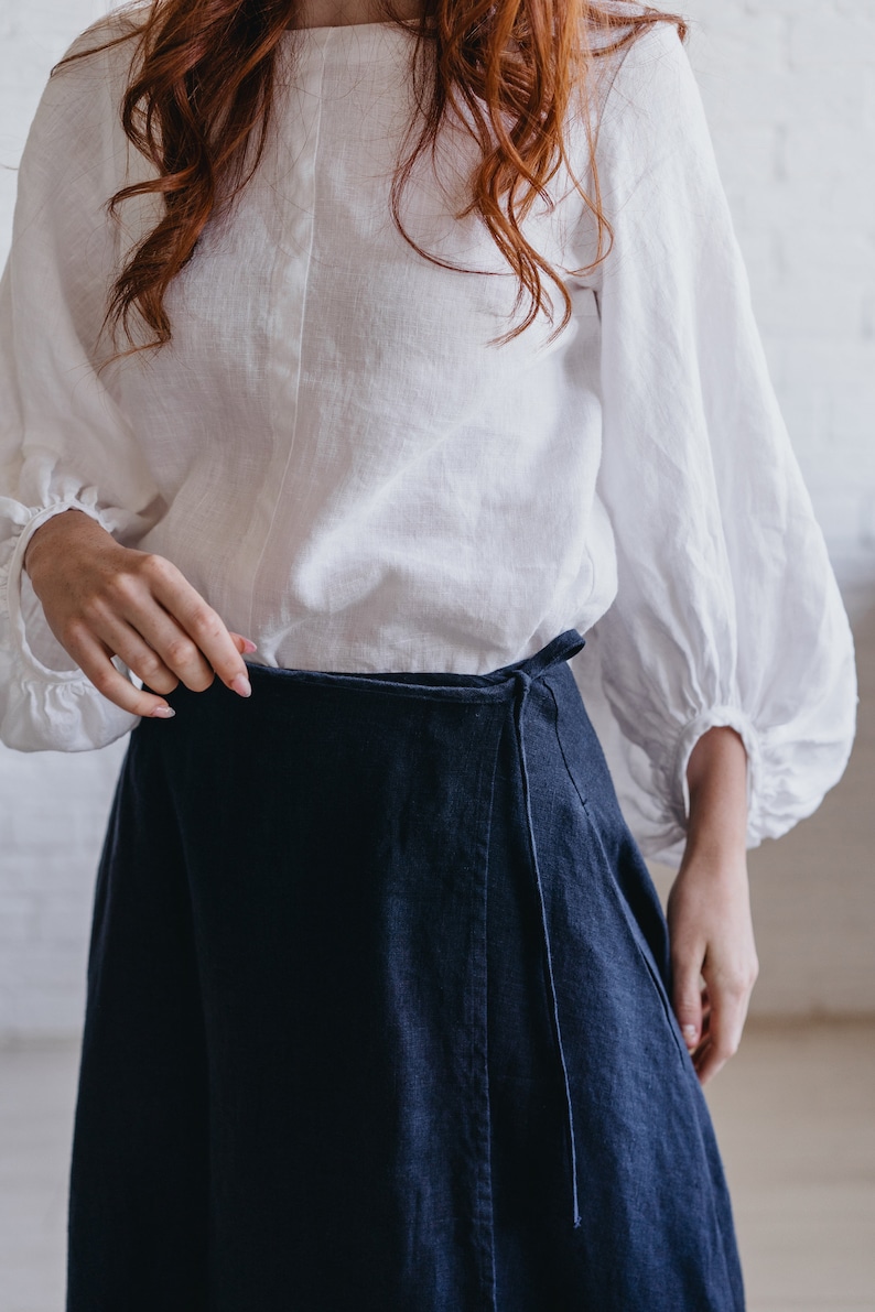 May include: A white linen blouse with long, gathered sleeves and a dark blue wrap skirt. The blouse has a simple neckline and the skirt ties at the waist. The outfit is made of natural fabrics.