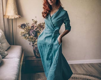 Könnte beinhalten: Ein türkisfarbenes Wickelkleid mit Bindegürtel und Taschen. Das Kleid hat Dreiviertelärmel und einen fließenden Rock. Die Frau trägt braune Sandalen und steht auf einem runden Jute-Teppich. Der Hintergrund umfasst ein Sofa, eine Lampe und eine Blumenvase.