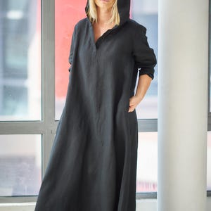 Peut inclure: Longue robe noire à capuche avec un col en V et des manches longues. La robe a une silhouette ample et fluide et des poches. La personne porte des lunettes de soleil. L'arrière-plan comprend une fenêtre et un pilier blanc.
