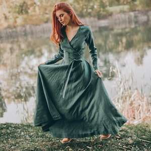 May include: A long, flowing, dark green dress with long sleeves and a wrap-style bodice. The dress is cinched at the waist with a matching fabric belt. The model is standing near a lake, wearing brown shoes.