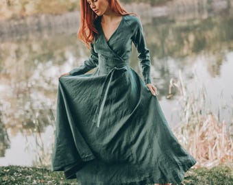 May include: A long, flowing, dark green dress with long sleeves and a wrap-style bodice. The dress is cinched at the waist with a matching fabric belt. The model is standing near a lake, wearing brown shoes.
