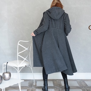 May include: A long, gray hooded coat with a flowing design. The coat is worn by a person standing in a room with a white chair and a white table.