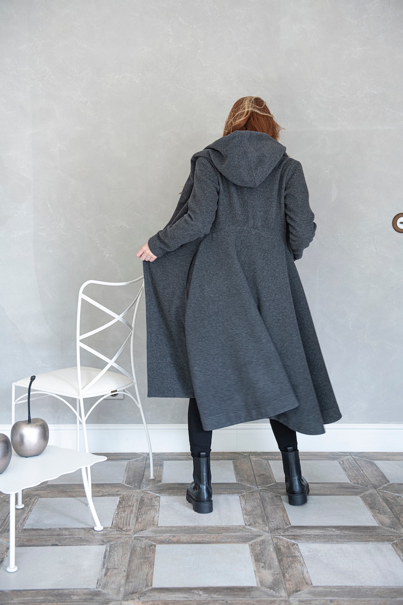 May include: A long, gray hooded coat with a flowing design. The coat is worn by a person standing in a room with a white chair and a white table.