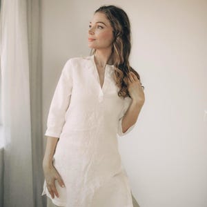 Op de afbeelding: Een witte linnen tuniek met een V-hals en driekwart mouwen. De tuniek wordt gecombineerd met lichtgekleurde broeken. De outfit is gemaakt van een lichte, ademende stof, perfect voor warm weer. De tuniek heeft een relaxte pasvorm en een eenvoudig, elegant ontwerp.