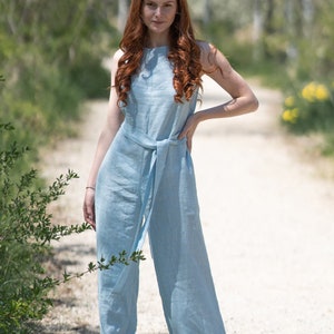 May include: A woman wearing a light blue linen jumpsuit with a wide belt. She is standing on a dirt path with trees in the background.