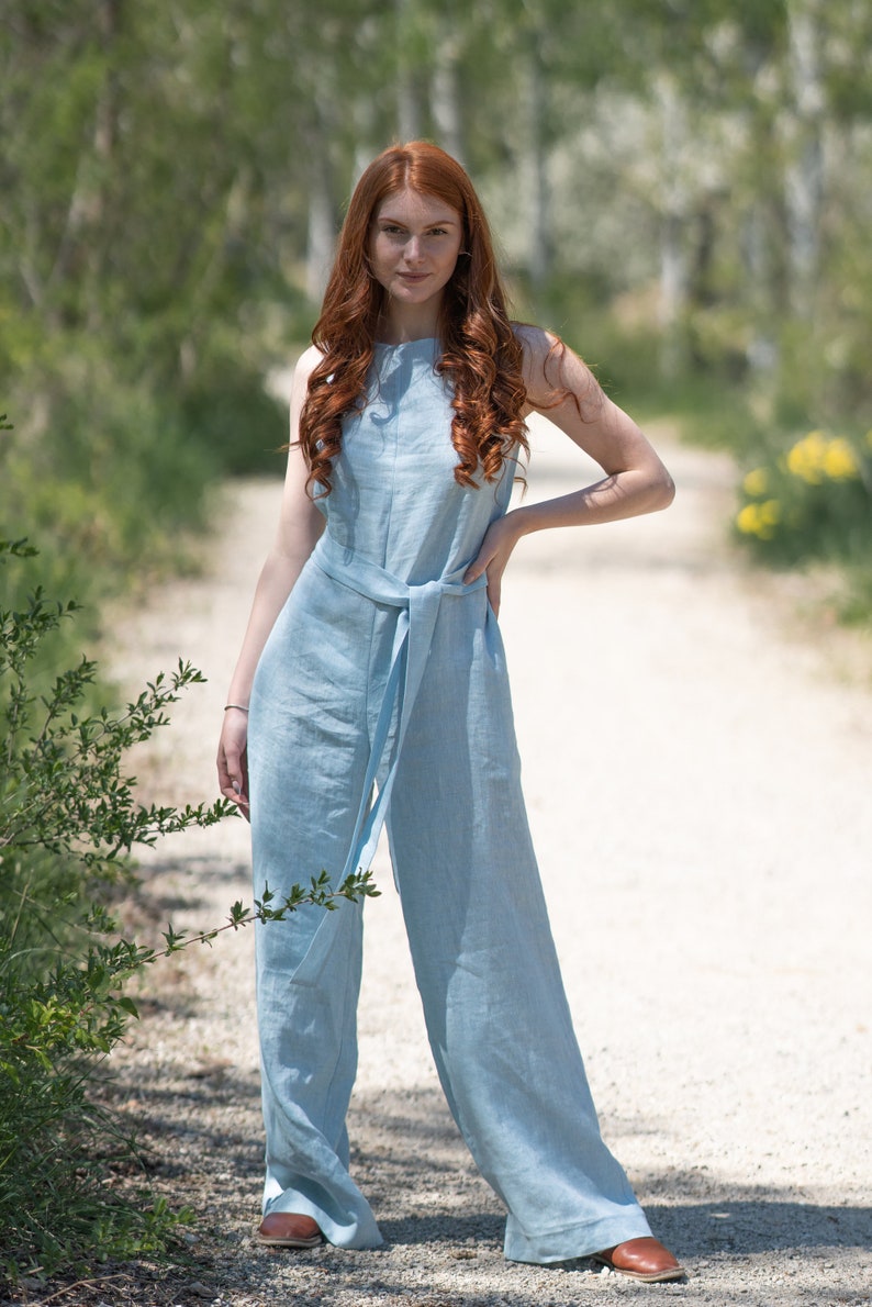 May include: A woman wearing a light blue linen jumpsuit with a wide belt. She is standing on a dirt path with trees in the background.