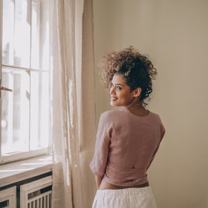 May include: A woman wearing a dusty rose wrap top and white linen pants stands near a window with a sheer curtain. The top has a wrap design, and the pants are wide-legged. The room has a neutral color palette.