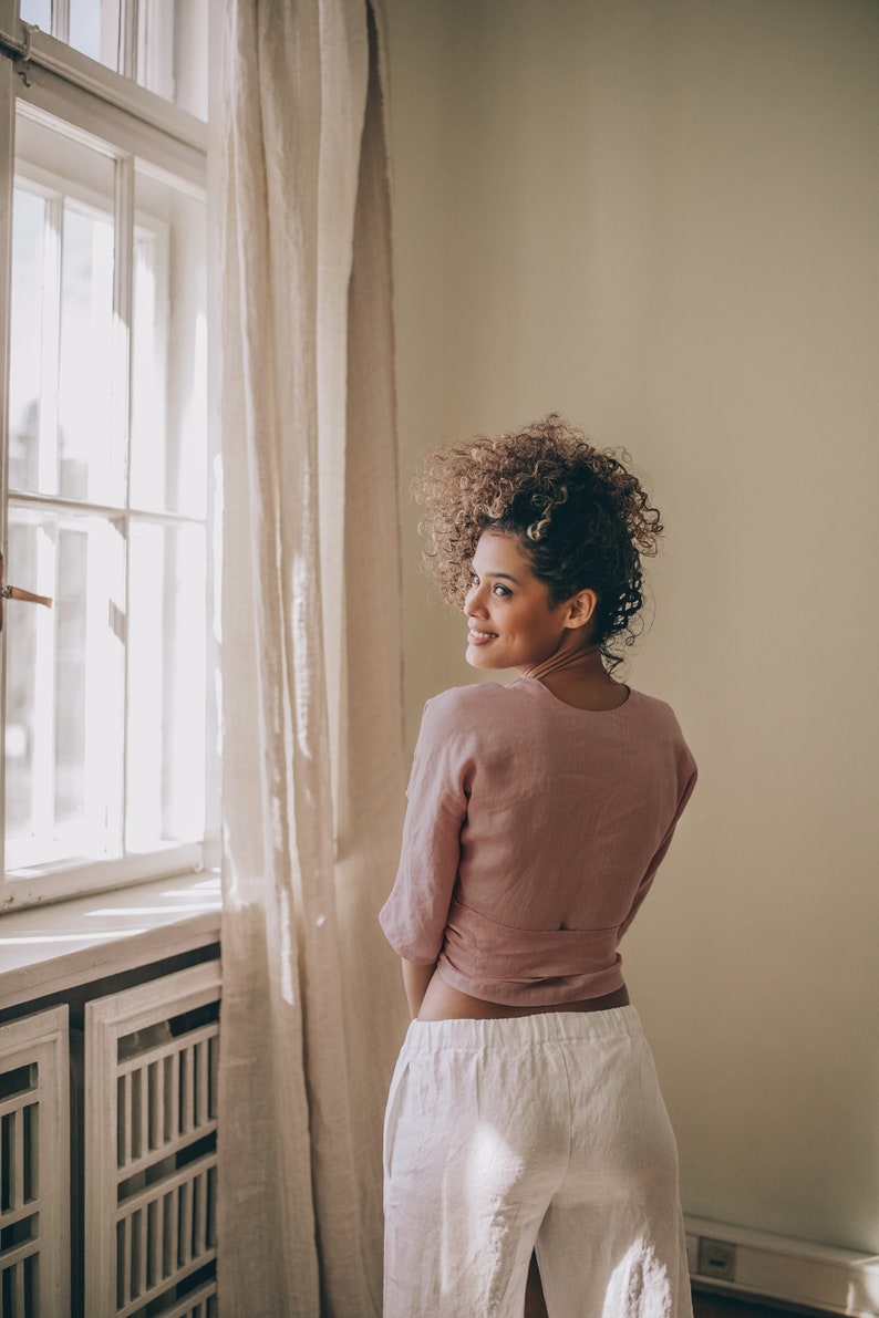 May include: A woman wearing a dusty rose wrap top and white linen pants stands near a window with a sheer curtain. The top has a wrap design, and the pants are wide-legged. The room has a neutral color palette.