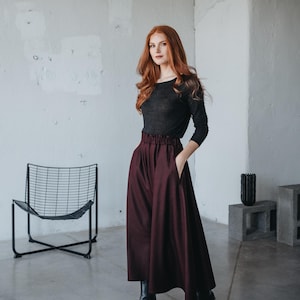 May include: A woman with long red hair wearing a black long-sleeved shirt and a burgundy high-waisted skirt. She is standing in a room with a white wall and a black metal chair.