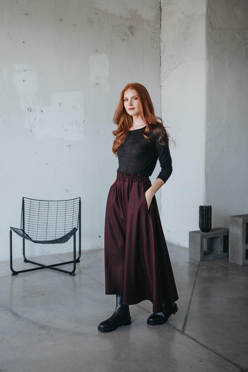 May include: A woman with long red hair wearing a black long-sleeved shirt and a burgundy high-waisted skirt. She is standing in a room with a white wall and a black metal chair.