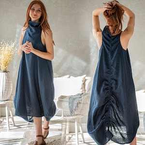 May include: A navy blue sleeveless dress with a high neckline. The dress has a unique, gathered design at the back and a bubble hem. The model is wearing brown sandals. The dress is made of a soft, flowing fabric.