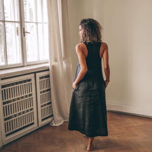 May include: A black sleeveless dress with a long, flowing skirt. The dress has pockets and is worn by a person standing near a window with a white frame and beige curtains. The room has hardwood floors and a white radiator.