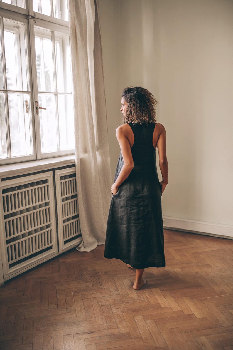 May include: A black sleeveless dress with a long, flowing skirt. The dress has pockets and is worn by a person standing near a window with a white frame and beige curtains. The room has hardwood floors and a white radiator.