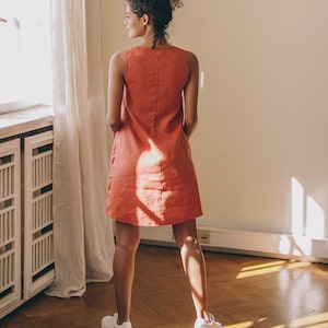 May include: A woman wearing a sleeveless, coral-coloured linen dress and white trainers. She is standing in a room with a window and wooden floors.